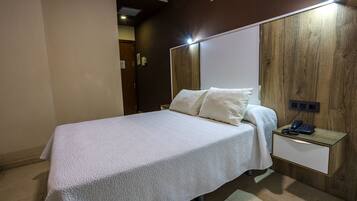 Una cama bien hecha con una colcha blanca, una mesita de noche con una lámpara y un cabecero de madera.