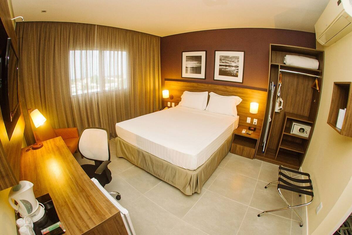 A hotel room with a bed, desk, chair, and a small table with a kettle. There are two wall-mounted lamps, a coat rack, and a wardrobe.