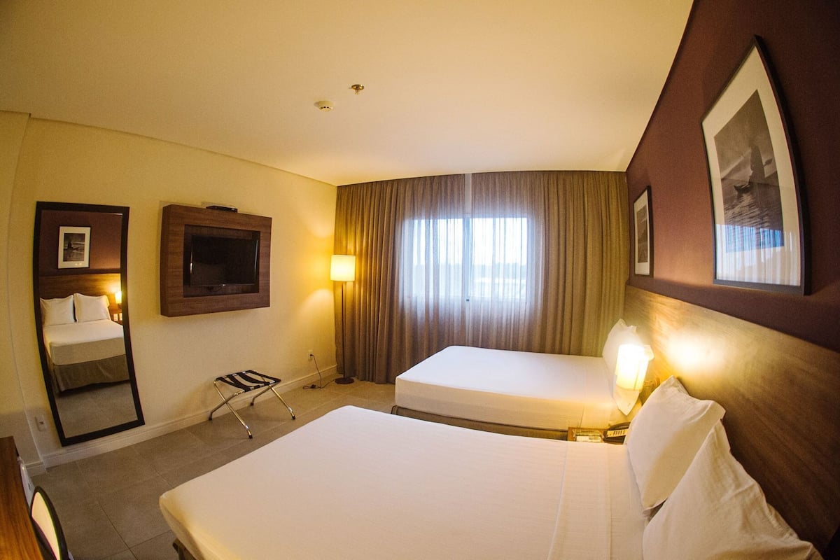 A hotel room with two beds, a TV, a mirror, a lamp, and a window with curtains.