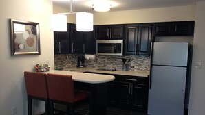 A compact kitchen with dark cabinets, a microwave, a refrigerator, and a small island with two bar stools. - Staybridge Suites Schererville by IHG (Schererville)