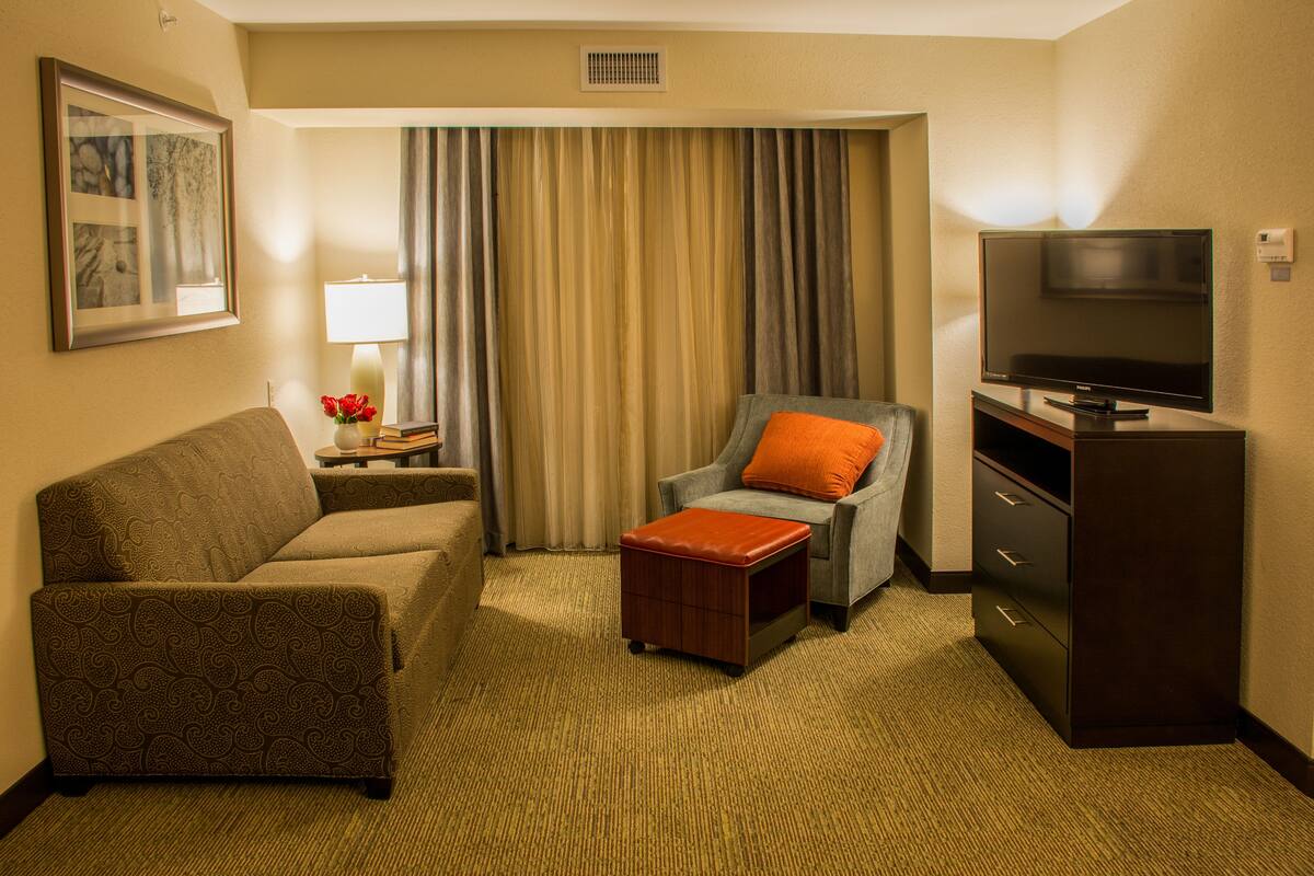 Hotelkamer met een bank, fauteuil, poef, televisie en een nachtkastje met lamp en bloemen.