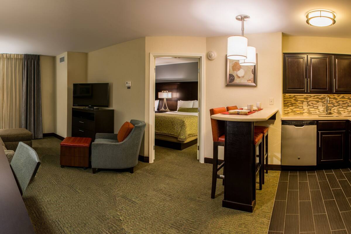 Een hotelkamer met een bureau, een stoel, een bank, een televisie, een kitchenette en een badkamer.