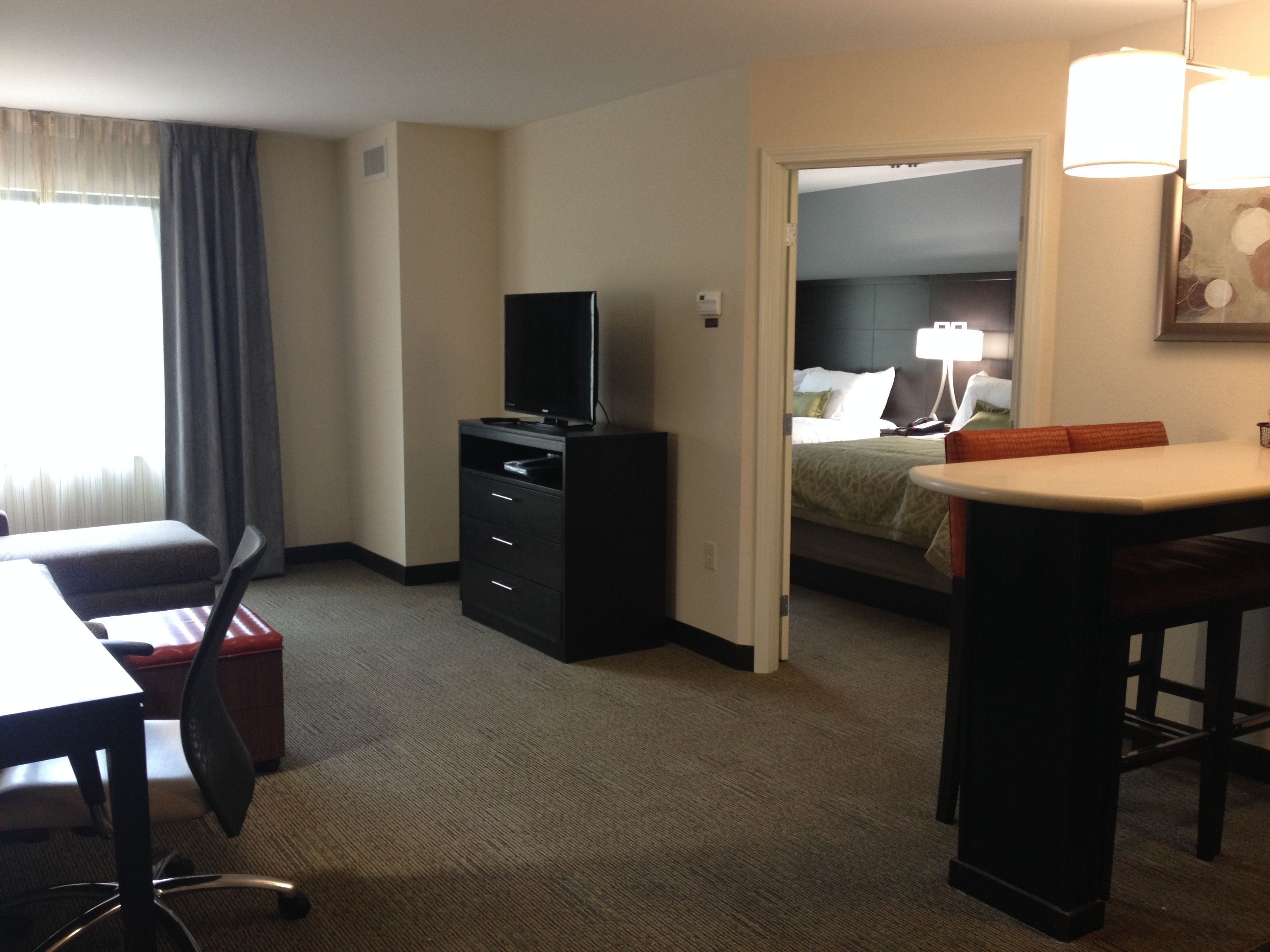 A hotel room with a desk, chair, TV, dresser, and a bed with a nightstand.