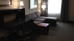 A hotel room with a desk, a sofa, a small ottoman, a chair, a lamp, a framed picture, and a window with curtains.