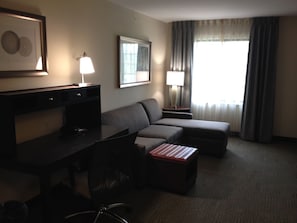 Een hotelkamer met een bureau, een bank, een klein voetenbankje, een stoel, een lamp, een lijst met een afbeelding en een raam met gordijnen.