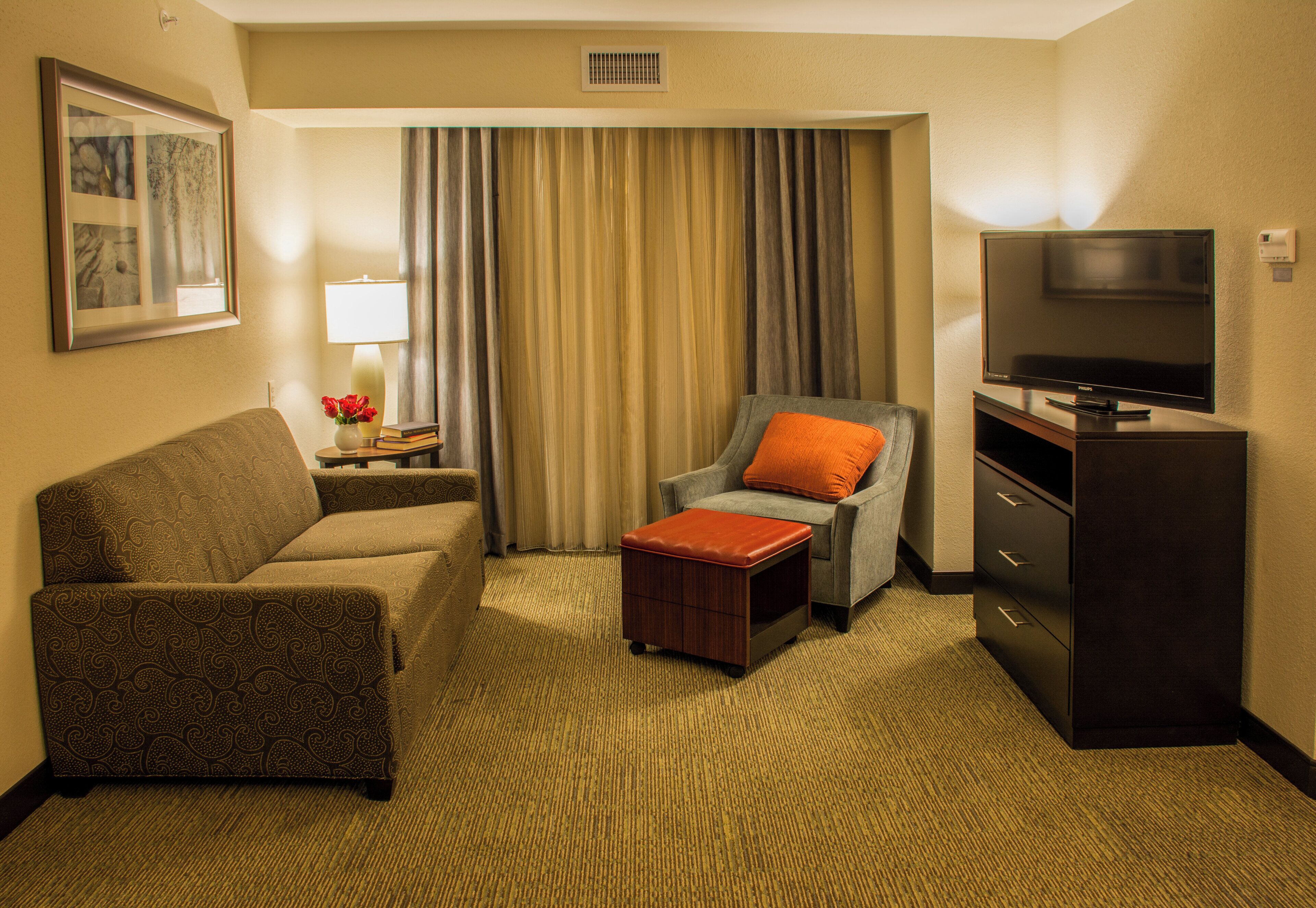 A hotel room with a sofa, armchair, ottoman, television, and a lamp on a side table.