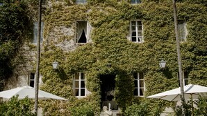 Un edificio con muros cubiertos de hiedra, una entrada central y mesas exteriores con sombrillas.