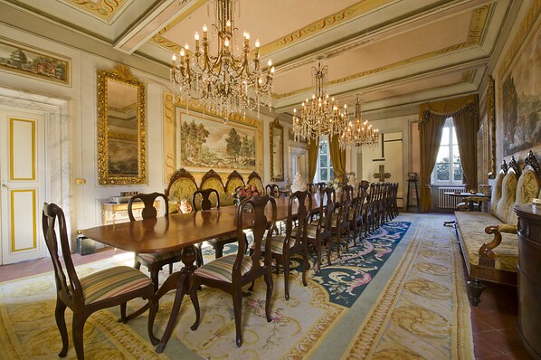 A formal dining room with a long wooden table, ornate chairs, chandeliers, and a large painting on the wall.
