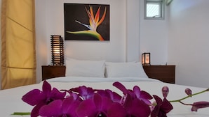 A neatly made bed with white linens and purple orchids placed on top. There is a painting of a bird of paradise on the wall behind the bed. - Baan Gecko (Krabi)
