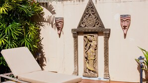 A sunlit outdoor lounge area with a beige cushioned lounge chair, a decorative wall with a relief sculpture, and a potted plant. - Baan Manu Chang (Krabi)