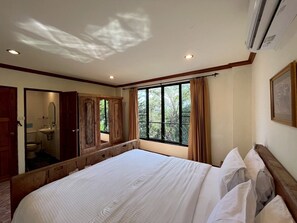 A bedroom with a large bed, a wooden wardrobe, a window with curtains, and a view of trees outside. - Thara Bayview Villa (Krabi)