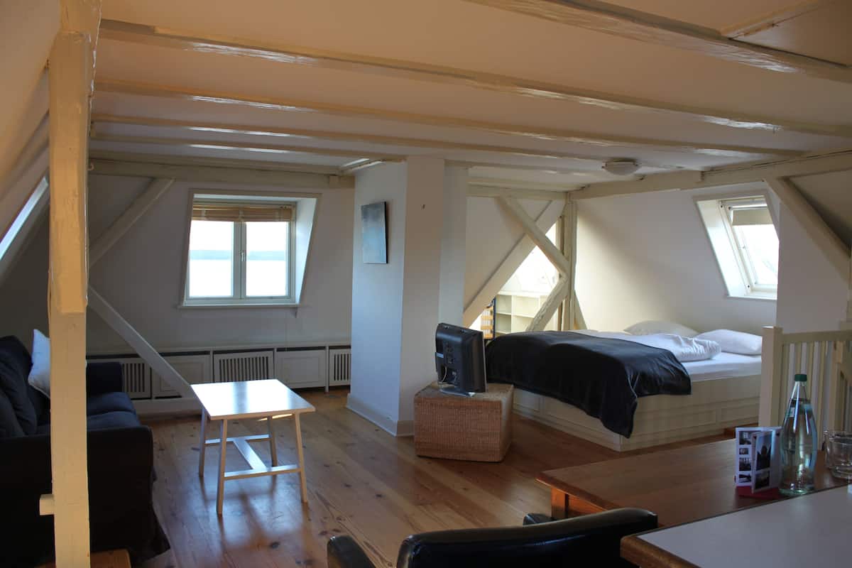 A loft-style bedroom with a bed, a small table, a TV, and a window.