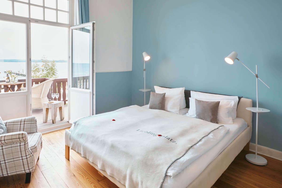 A bedroom with a bed, two bedside lamps, a view through a large window, and a balcony.