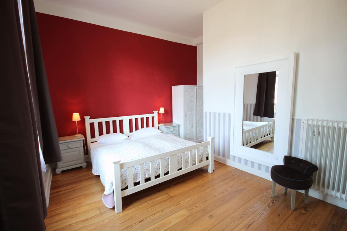 A bedroom with a wooden floor, a white bed with a headboard, a nightstand with a lamp, a wardrobe, and a mirror on the wall.