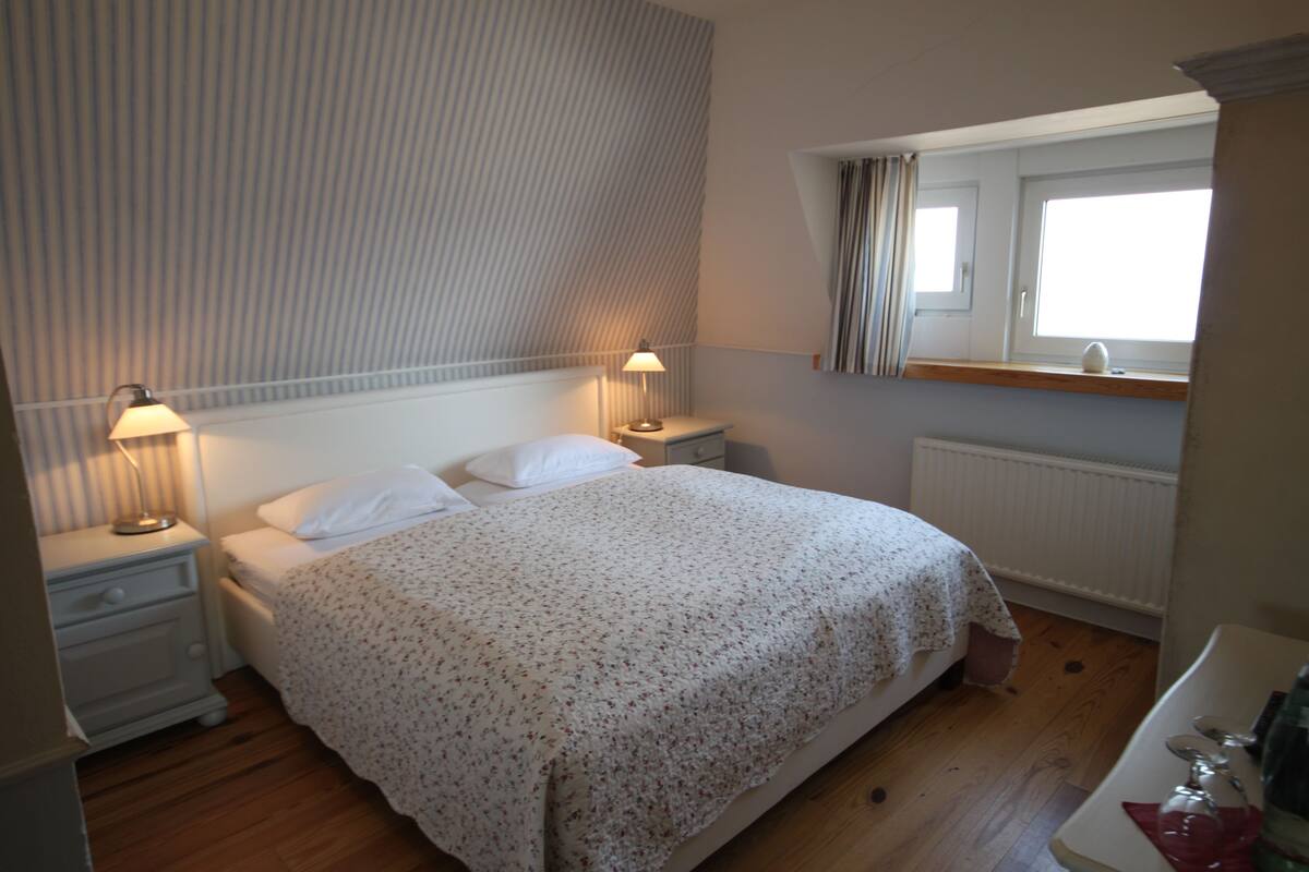 A bedroom with a bed, two bedside tables with lamps, a window with curtains, and a radiator.