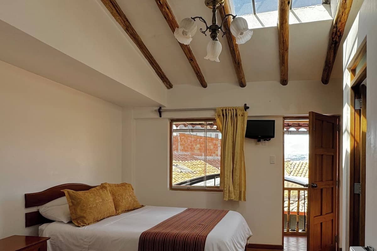 Quarto com duas camas, vigas de madeira no teto, uma janela com cortinas e uma TV fixada na parede.
