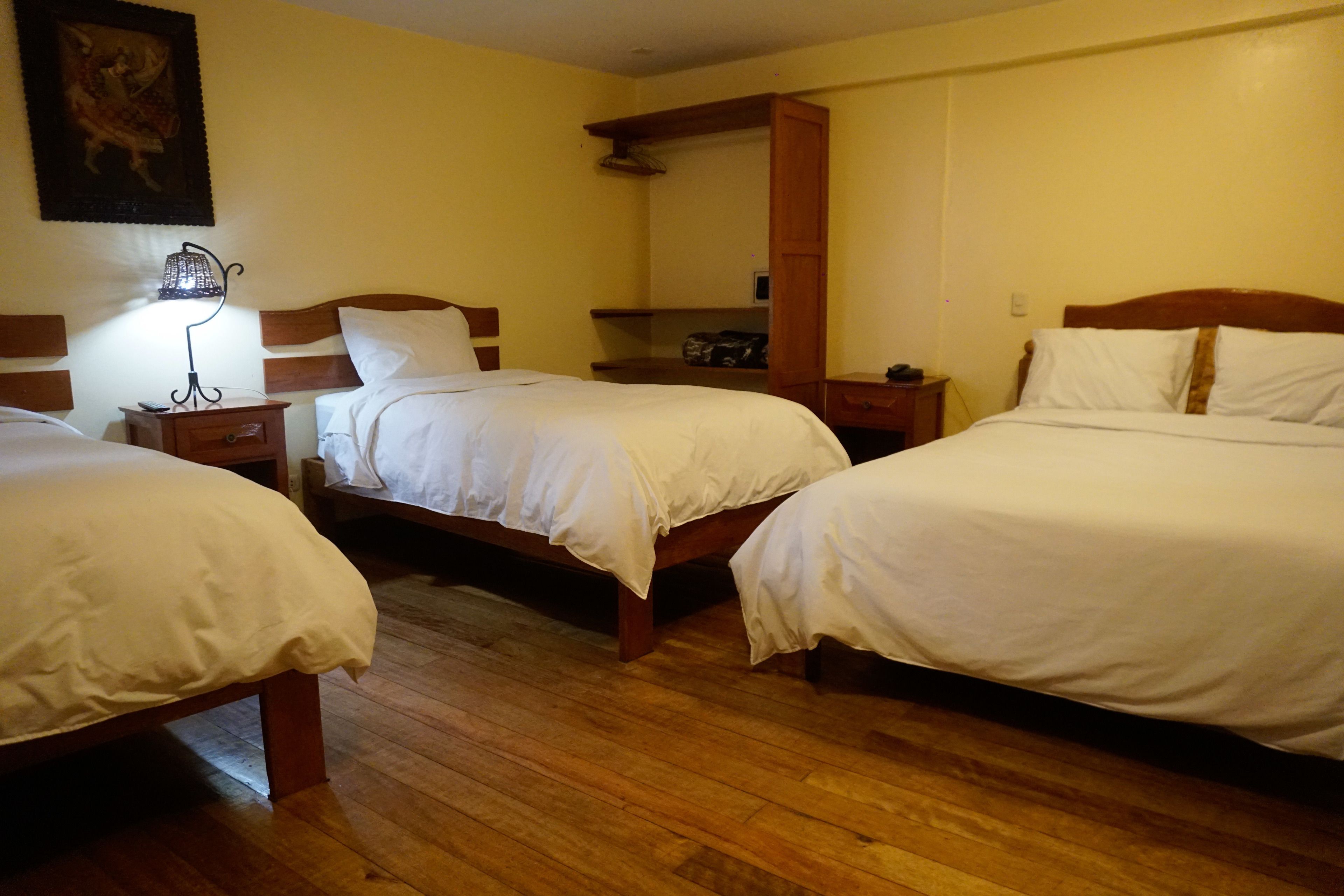A hotel room with three single beds, wooden floors, a nightstand with a lamp, and a painting on the wall.