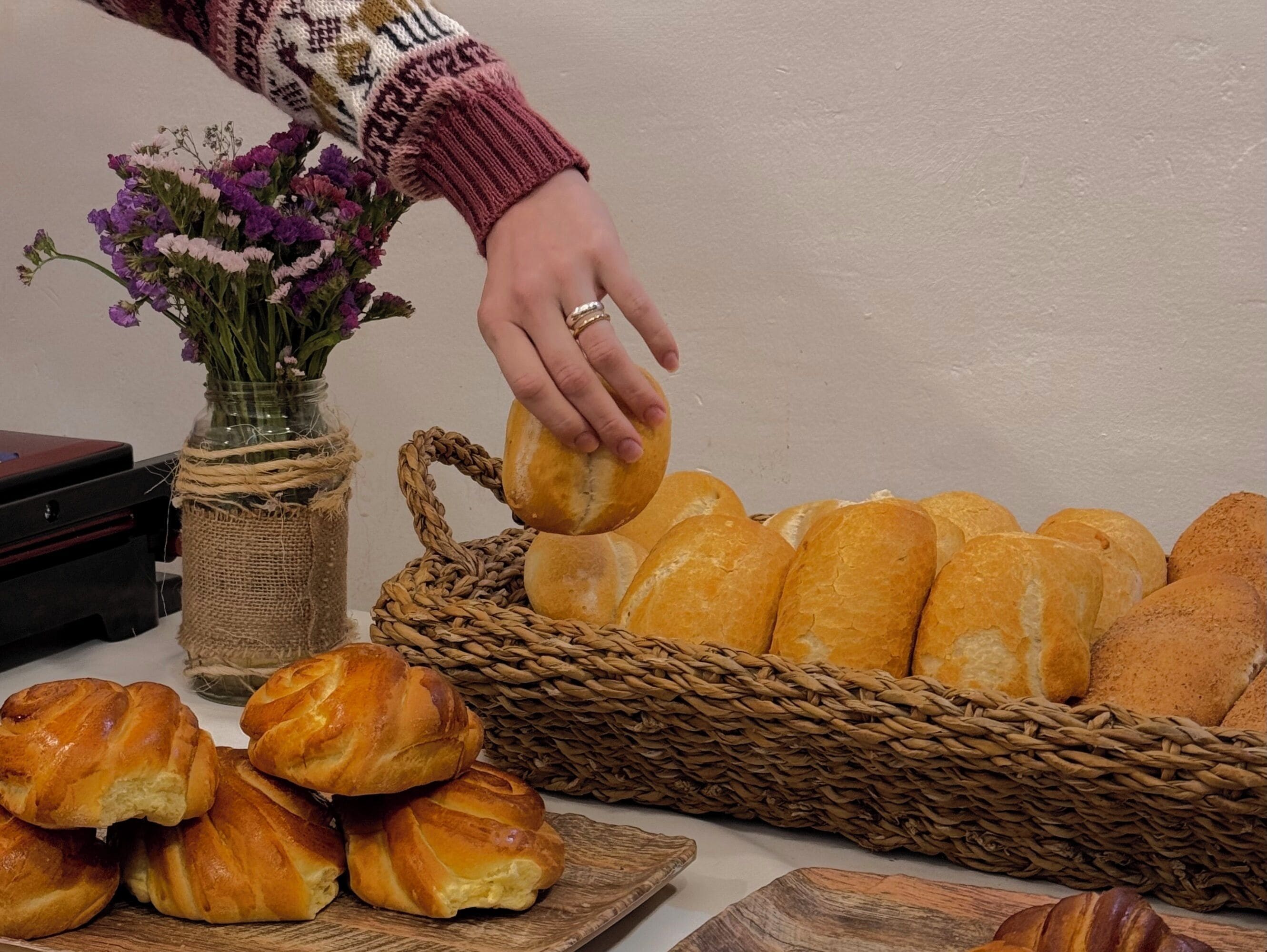 Una mano alcanzando una hogaza de pan de una cesta, con un jarrón de flores y una tabla de madera con cruasanes al frente.