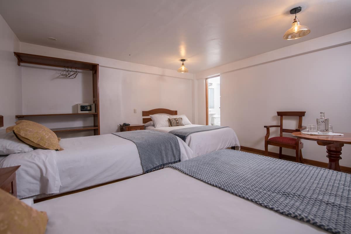 Quarto de hotel com duas camas, uma cabeceira de madeira, uma mesinha com uma cadeira, micro-ondas e uma janela.