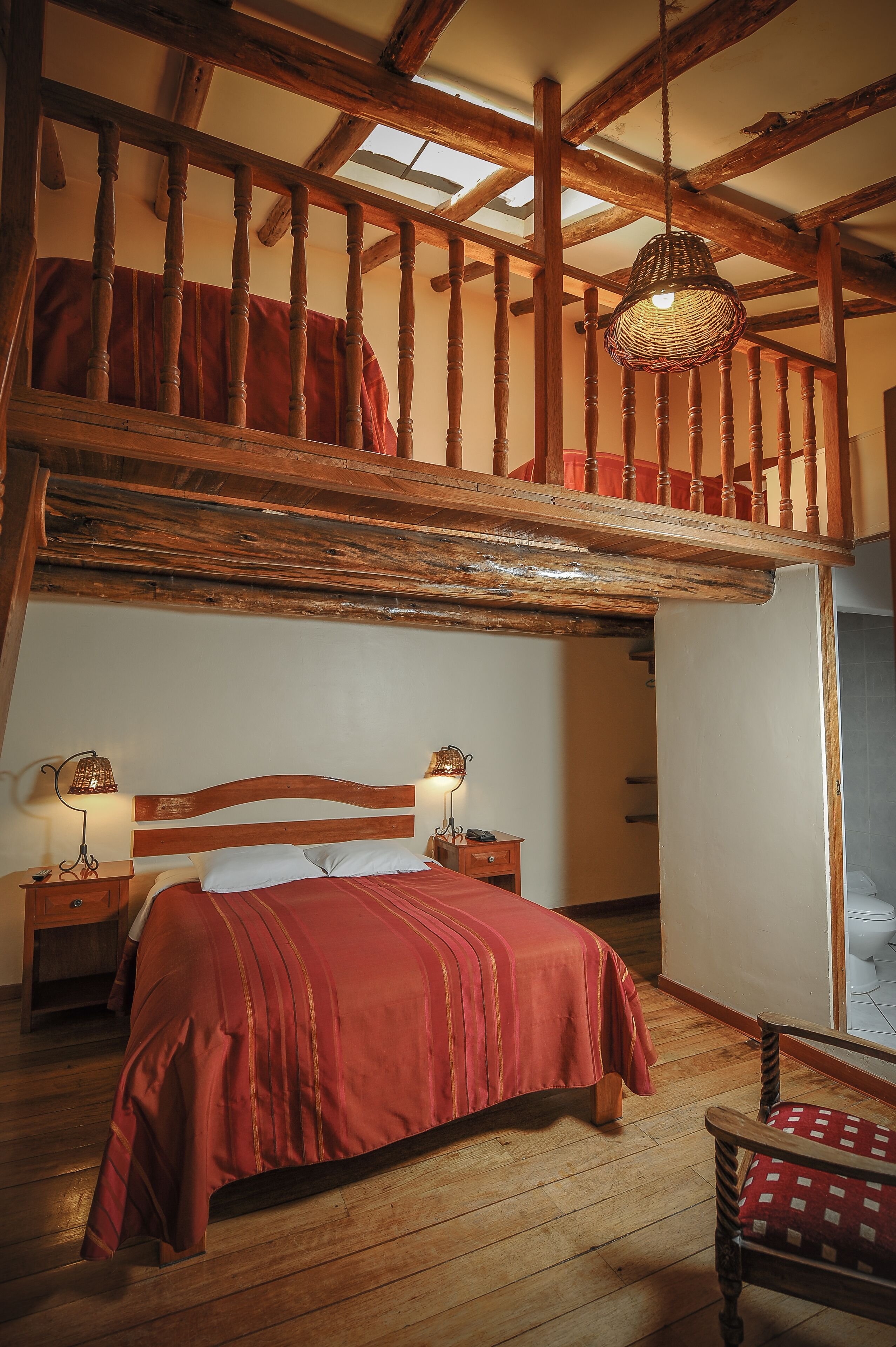 A bedroom with a wooden bed, two bedside tables with lamps, a red blanket, and a wooden chair.