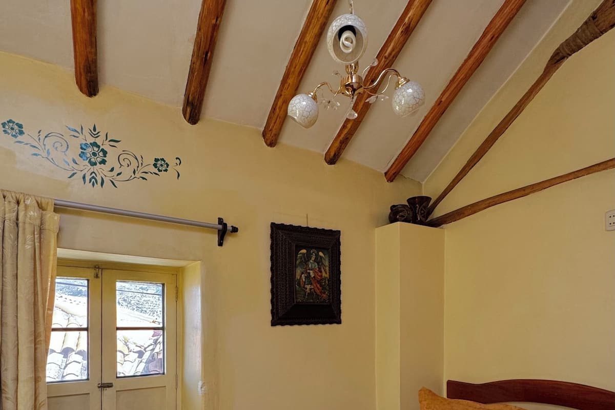 Quarto com teto de madeira, cama com colcha estampada, um quadro na parede e janela com cortinas.