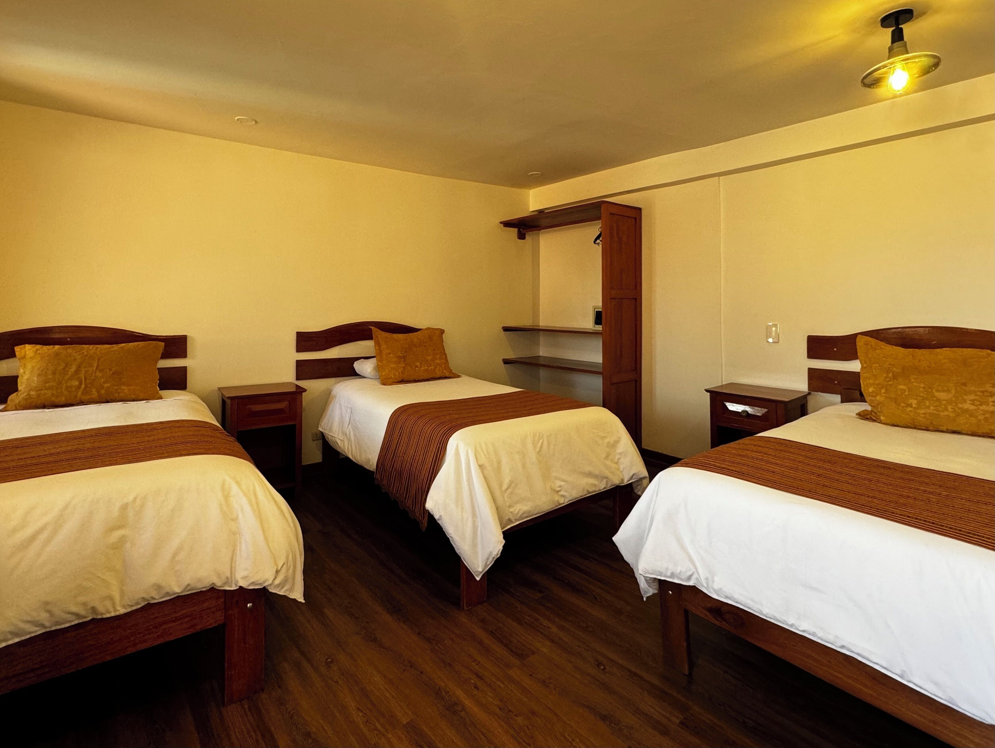 A hotel room with three wooden beds, each with a white and brown bedspread. There is a wooden nightstand with a lamp on each bed. The room has wooden flooring and a wooden ceiling with a light fixture.