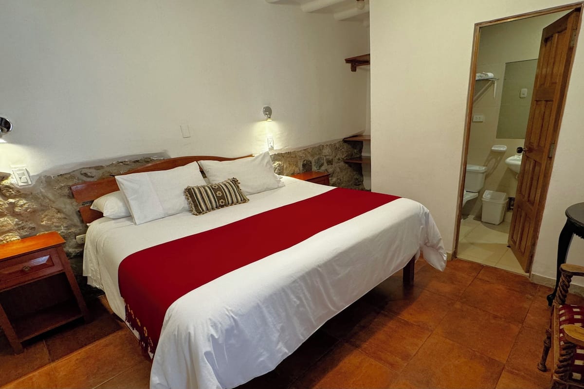 Quarto com cama, mesas de cabeceira, uma cadeira de madeira, uma pequena mesa com objetos e um banheiro visível por uma porta aberta.