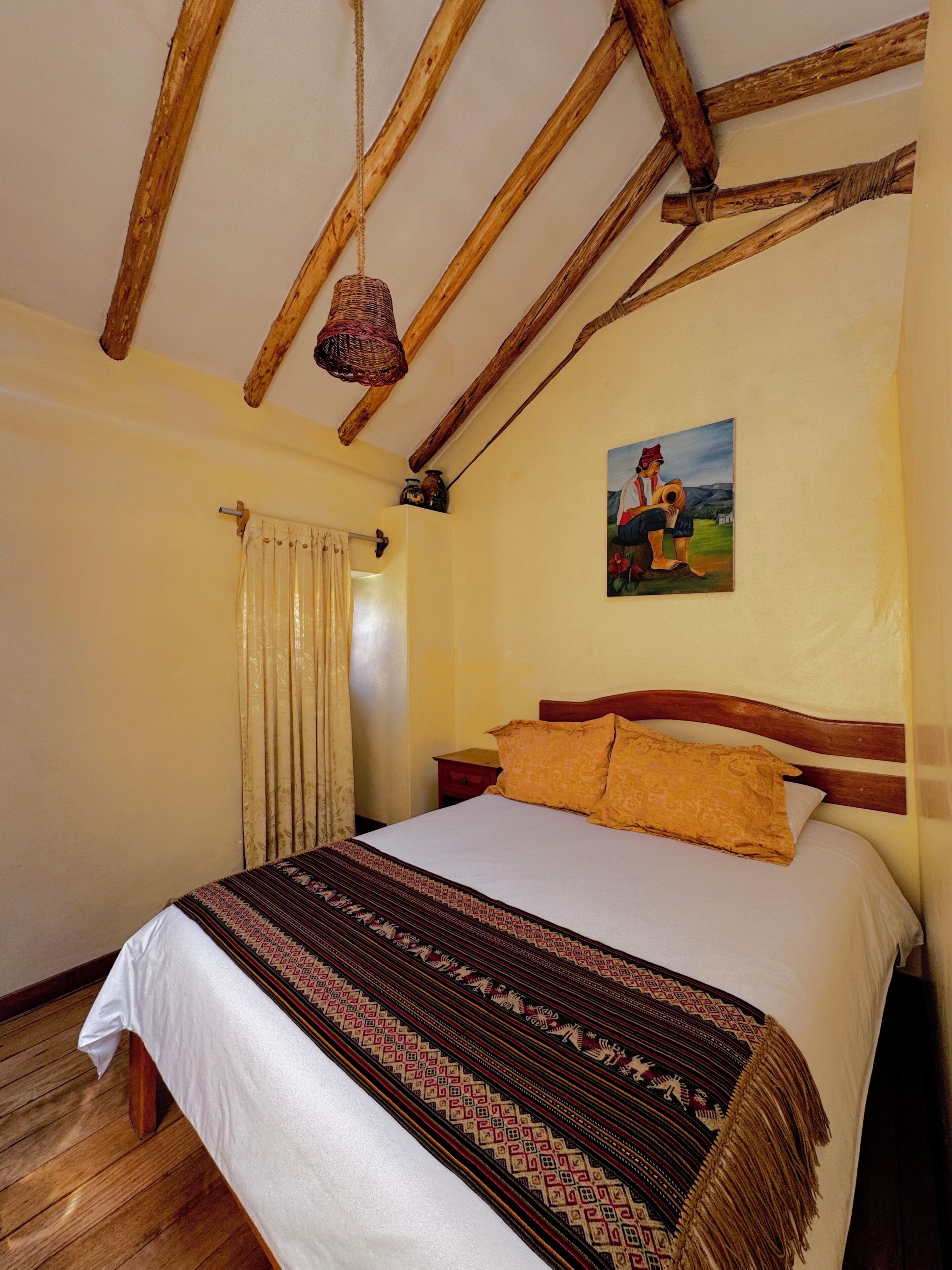 Ein Schlafzimmer mit Holzbalkendecke, einem Bett mit gemusterten Betttextilien, einem Bild an der Wand und einem Fenster mit Vorhängen.