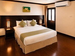 Une chambre d’hôtel avec une tête de lit en bois, un lit recouvert de draps blancs et agrémenté d’oreillers verts, une table de chevet avec une lampe et un appareil de climatisation fixé au mur.