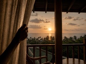 Ein Balkon mit Blick auf einen Sonnenuntergang über dem Meer, einem Holellentrail und einer Person, die hinter einem Vorhang teilweise zu sehen ist.