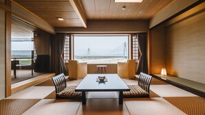 A traditional Japanese-style room with tatami flooring, a low table, and two chairs. A large window offers a view of a bridge and landscape.