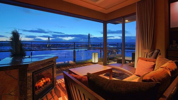 A modern living room with a fireplace, comfortable seating, and a large window offering a view of a bridge and water.