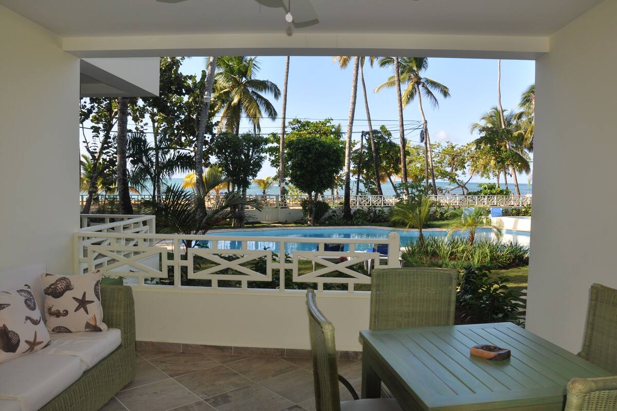 A balcony with a view of a pool, palm trees, and the ocean.