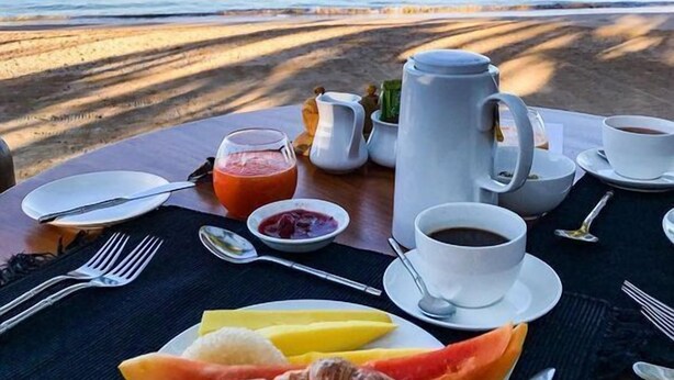 Disposição para o café da manhã em uma praia, com vista para o oceano, incluindo croissants, frutas, café e suco.