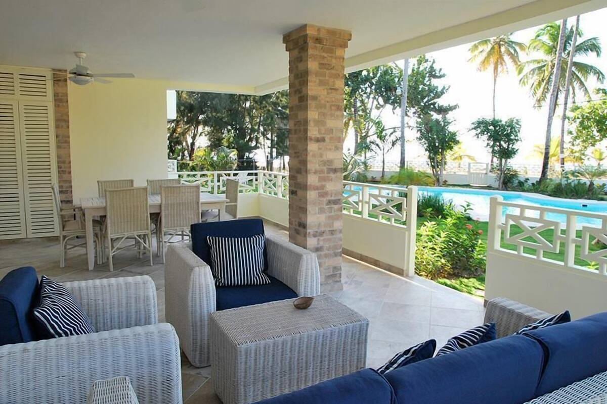 A covered outdoor area with wicker furniture, a pool, and palm trees.