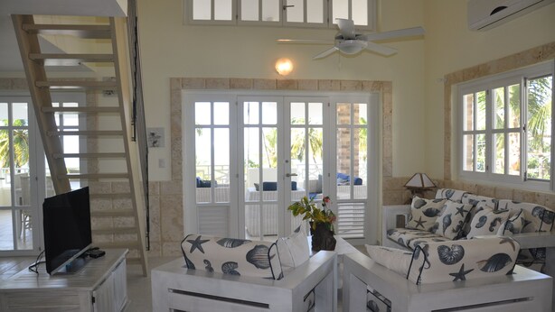 A spacious living area with a TV on a cabinet, white furniture with starfish patterns, a ceiling fan, and large windows with a view of palm trees. - Hotel Residence Marilar (Las Terrenas)