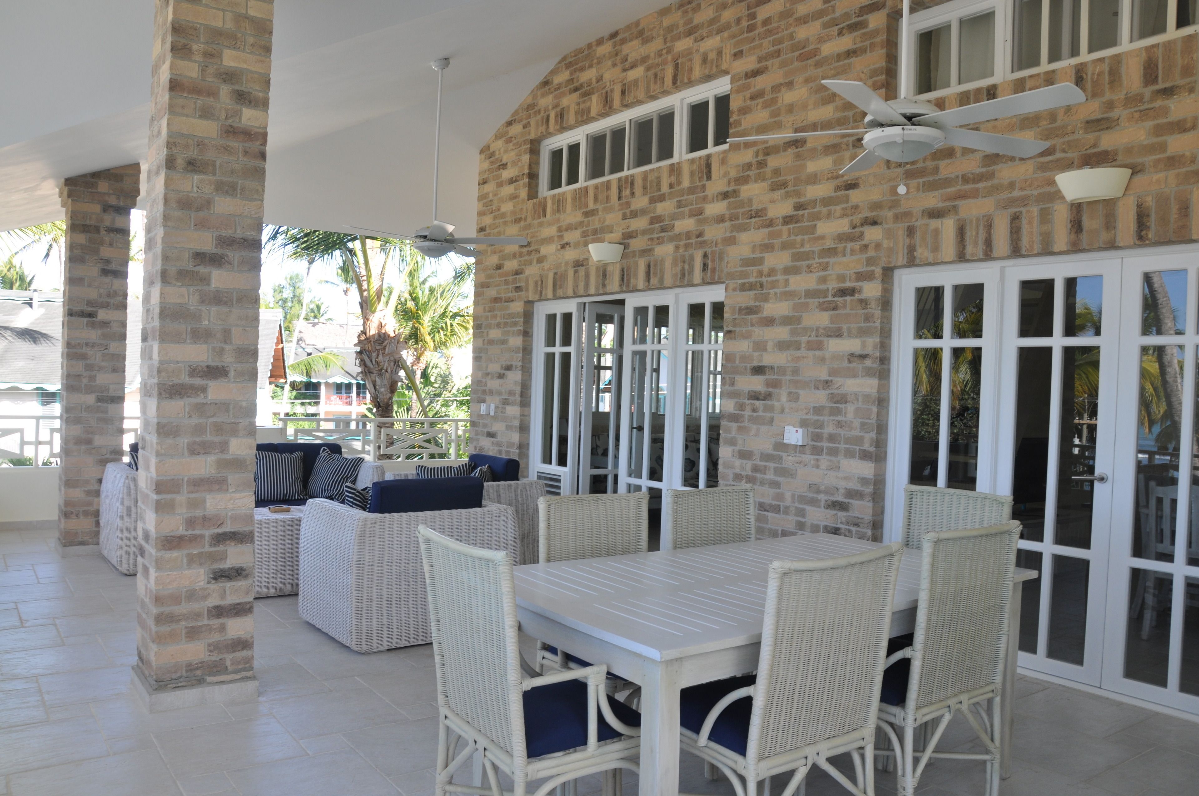 A covered patio area with a brick wall, white wicker furniture, and a ceiling fan.