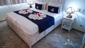 Cama bem arrumada com colcha azul e branca, duas almofadas e uma composição decorativa de toalhas. Tem uma mesa de cabeceira com abajur e um banquinho de madeira pequeno. O quarto tem piso de azulejo e cabeceira branca.