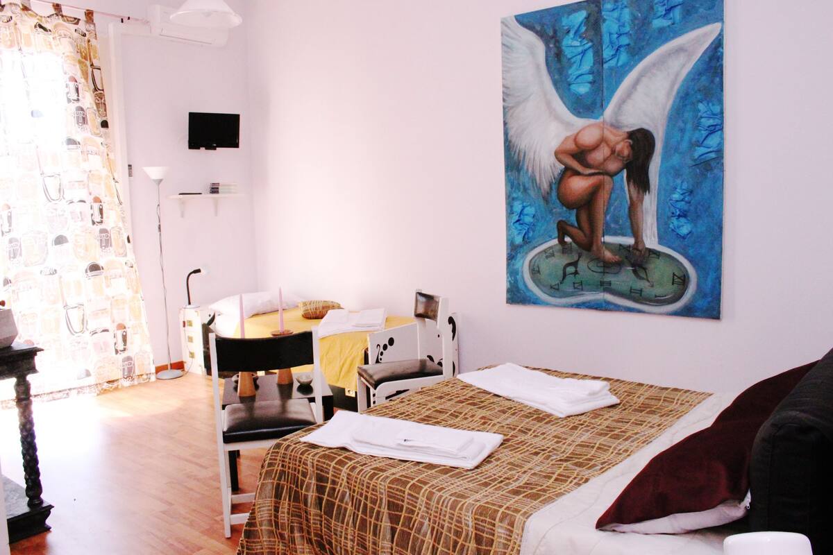 Quarto de hotel com cama, escrivaninha, cadeira, quadro na parede e janela com cortinas.