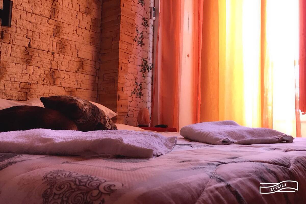 Uma cama com almofadas e um cobertor, uma parede de tijolos e uma janela com cortinas.