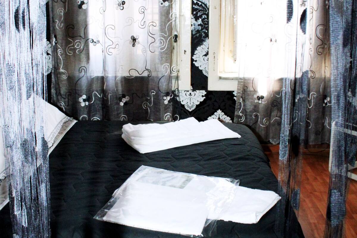 Quarto com cama coberta por roupa de cama preta, janela com cortinas brancas e chão de madeira.
