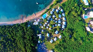 An aerial view of a resort with multiple bungalow-style accommodations clustered around a central pool area, surrounded by dense greenery and a beach. - Bungalows on the Bay (Christiansted)
