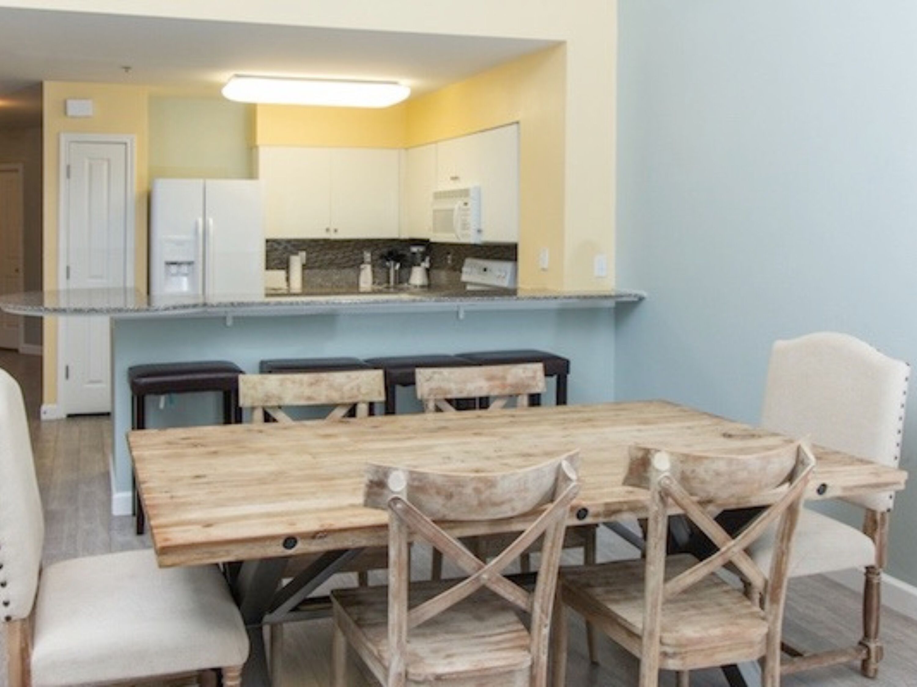 A dining area with a wooden table and chairs, a kitchen in the background, and a light blue wall.
