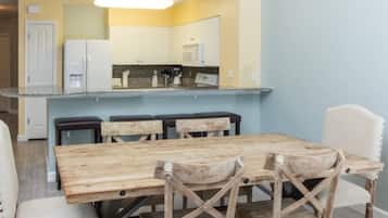 A dining area with a wooden table and chairs, a kitchen in the background, and a light blue wall.