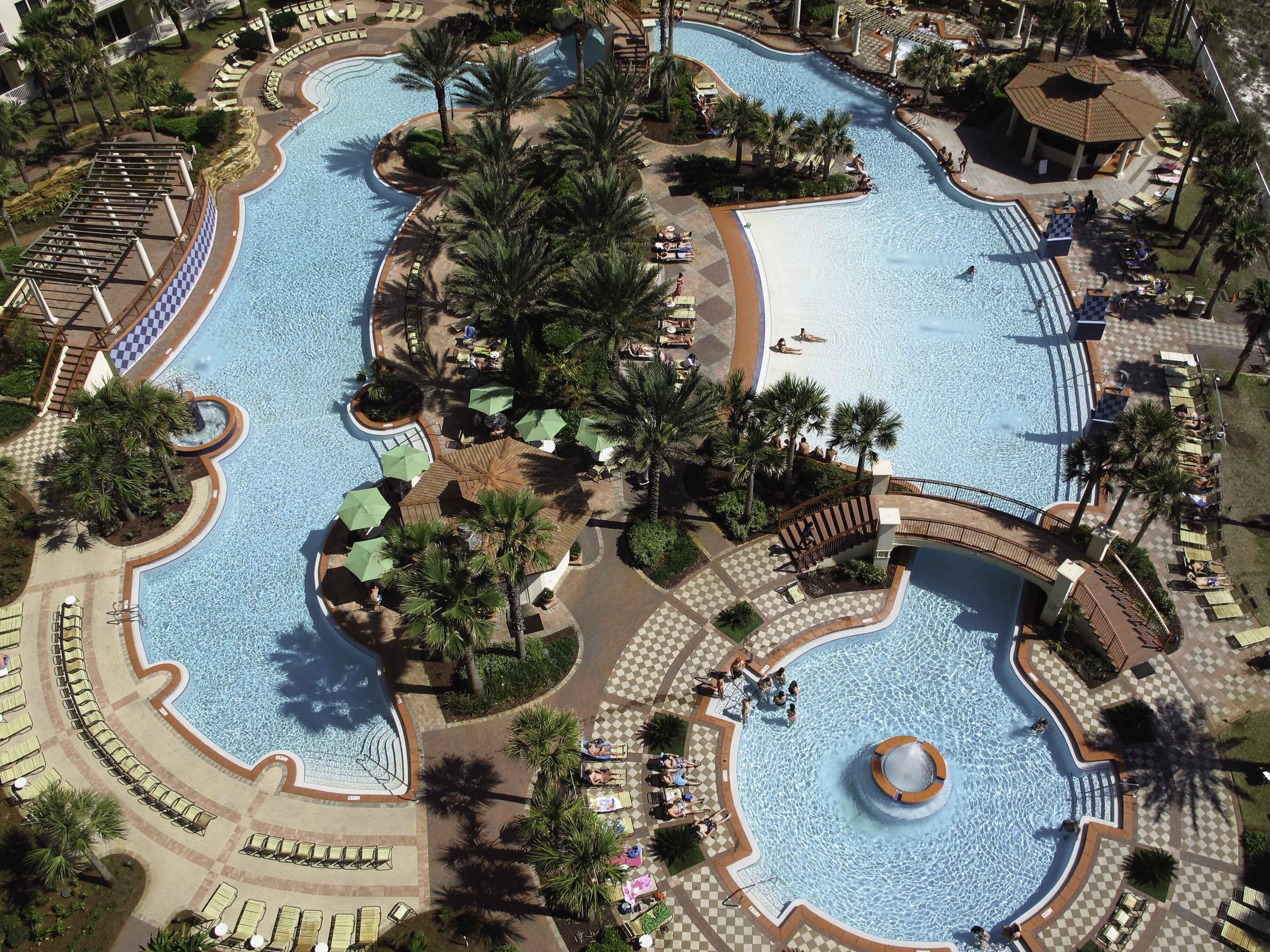 A large, multi-level swimming pool complex with various water features, surrounded by palm trees and sun loungers.