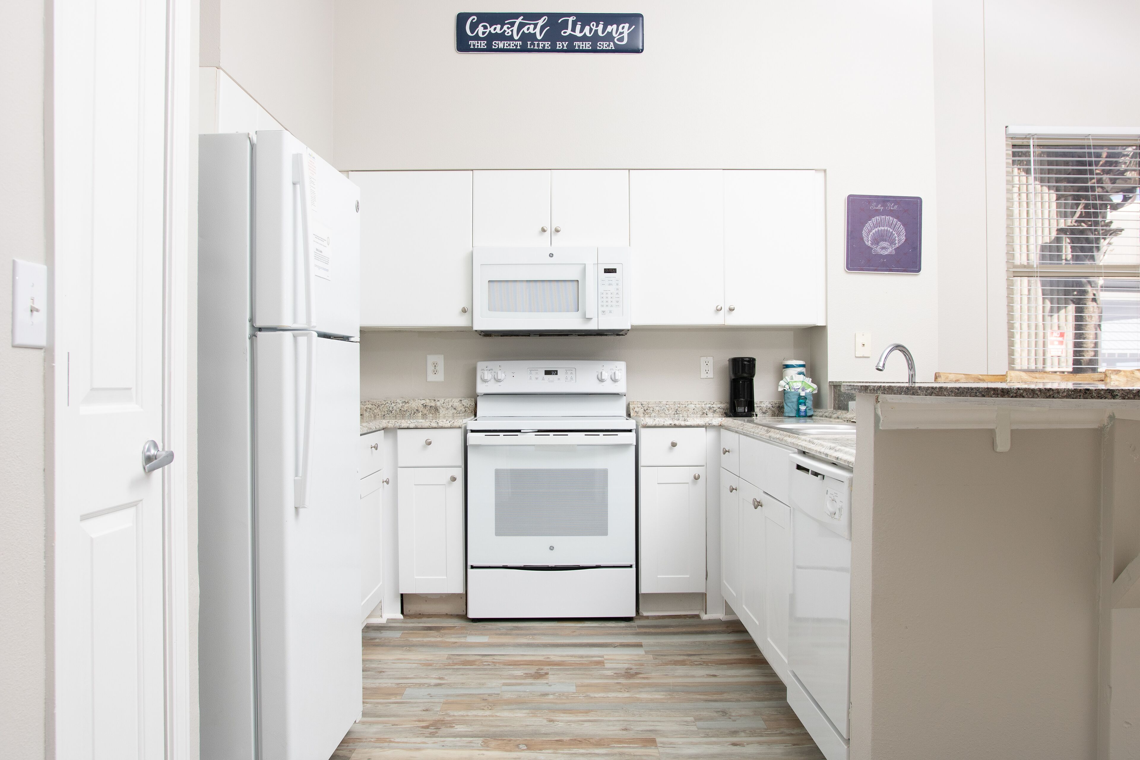 Una cocina moderna con armarios blancos, microondas integrado, horno y lavavajillas. Un cartel sobre el microondas dice 'Coastal Living'.