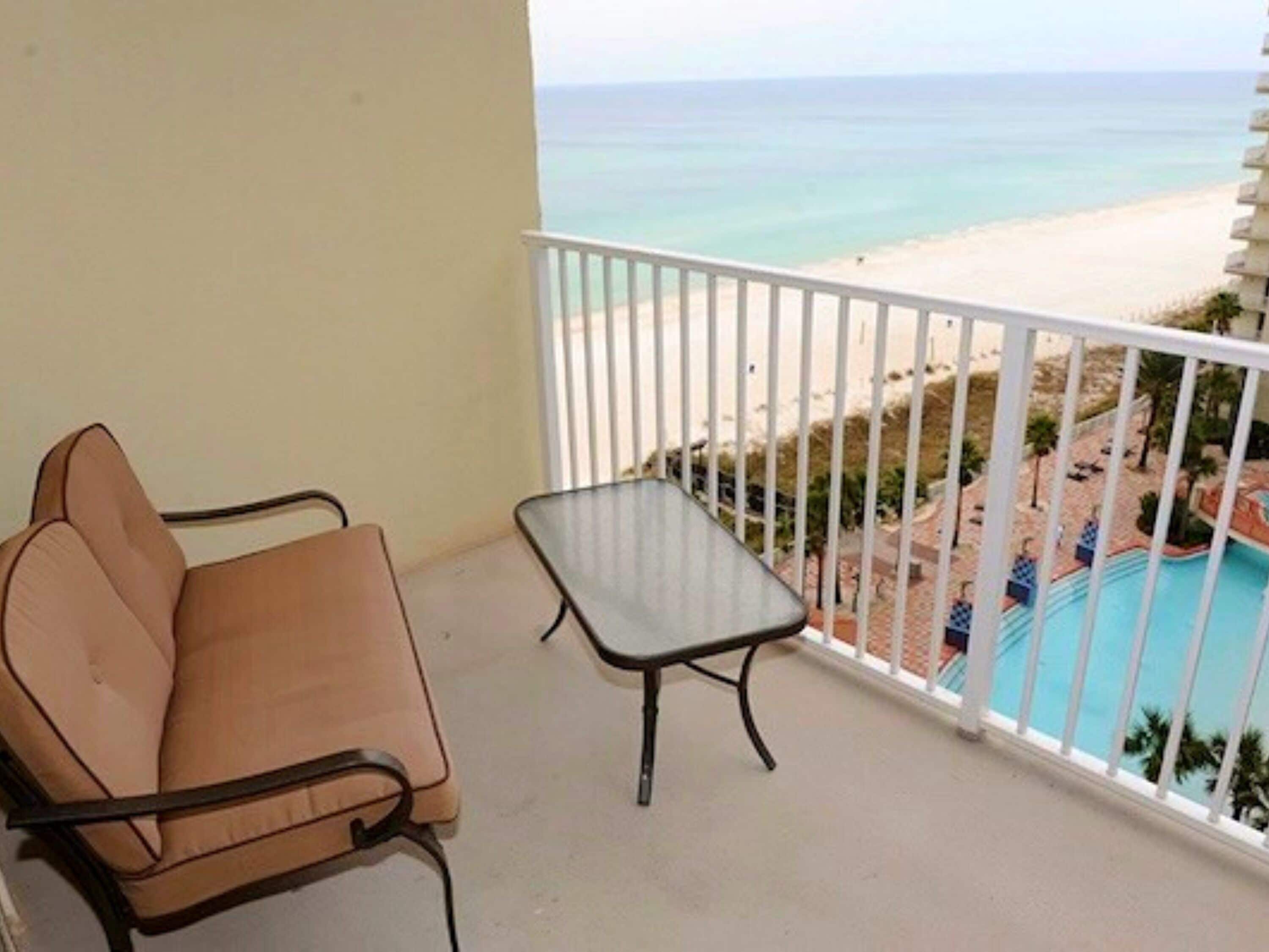 Un balcón con un sofá color crema y una mesita pequeña, con vistas a una playa y una piscina.