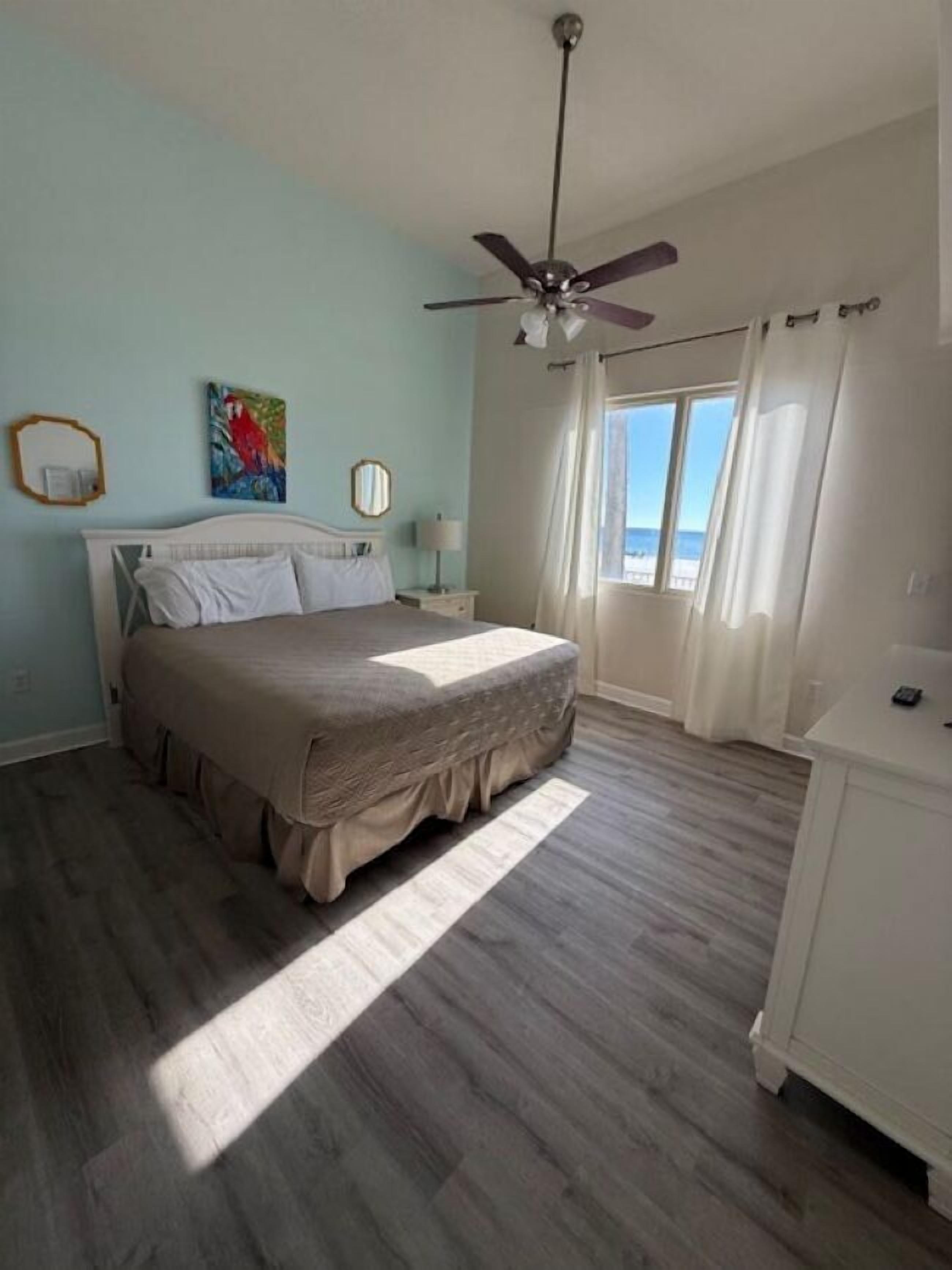 Een slaapkamer met een bed, een plafondventilator, een raam met gordijnen en uitzicht op de oceaan.