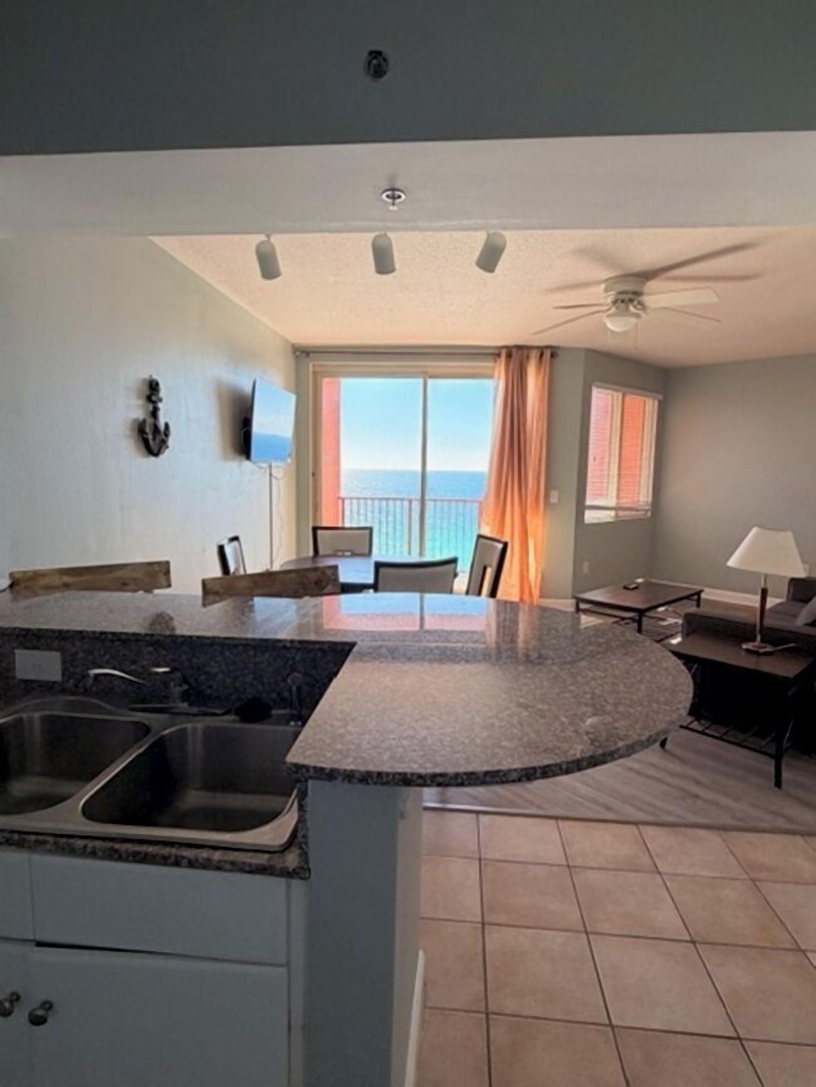 Cocina compacta con una isla central, comedor con mesa y sillas, ventilador de techo y vistas al océano a través de amplias ventanas.