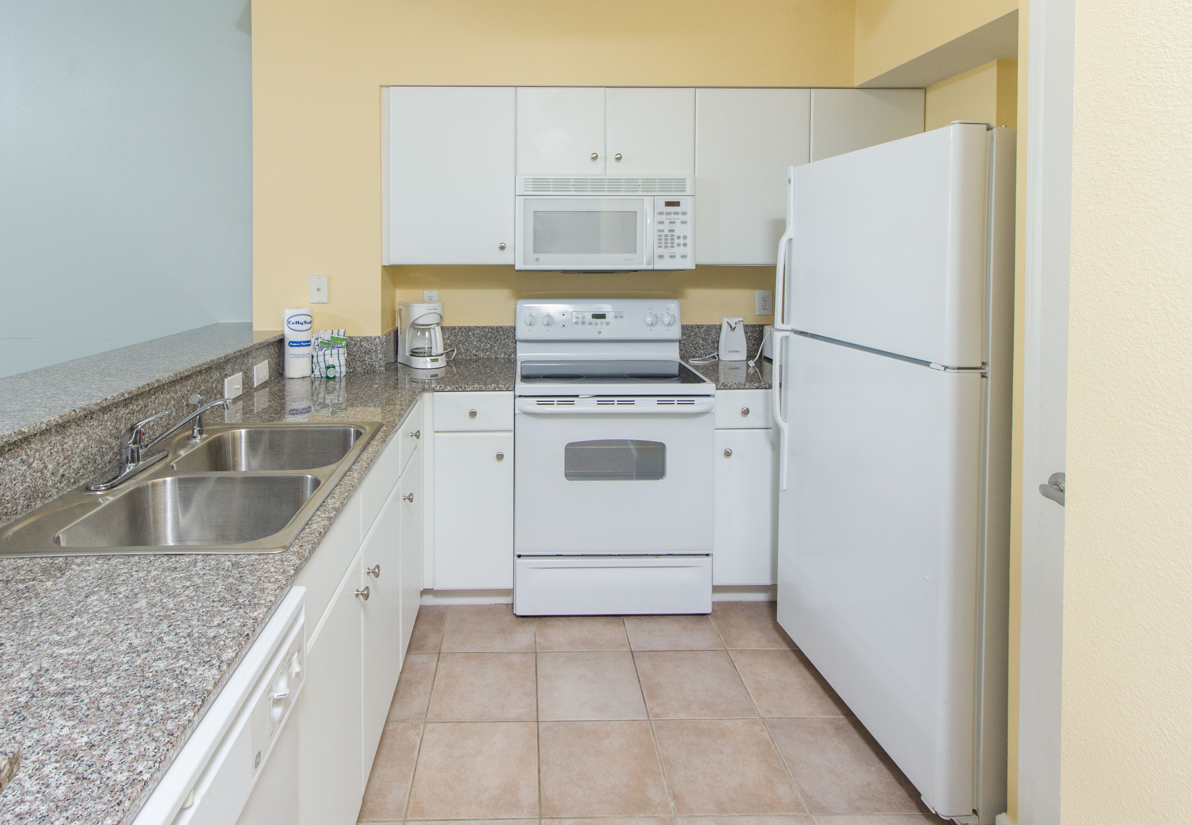 Una cocina compacta con electrodomésticos blancos, encimeras de granito y baldosas color beige.